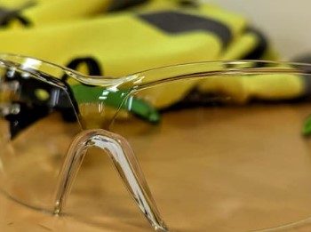 Clear safety goggles placed on a wooden table with a yellow high-visibility vest blurred in the background.
