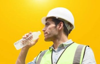 Construction worker in a high-vis vest and hard hat drinking water from a bottle against a bright yellow background